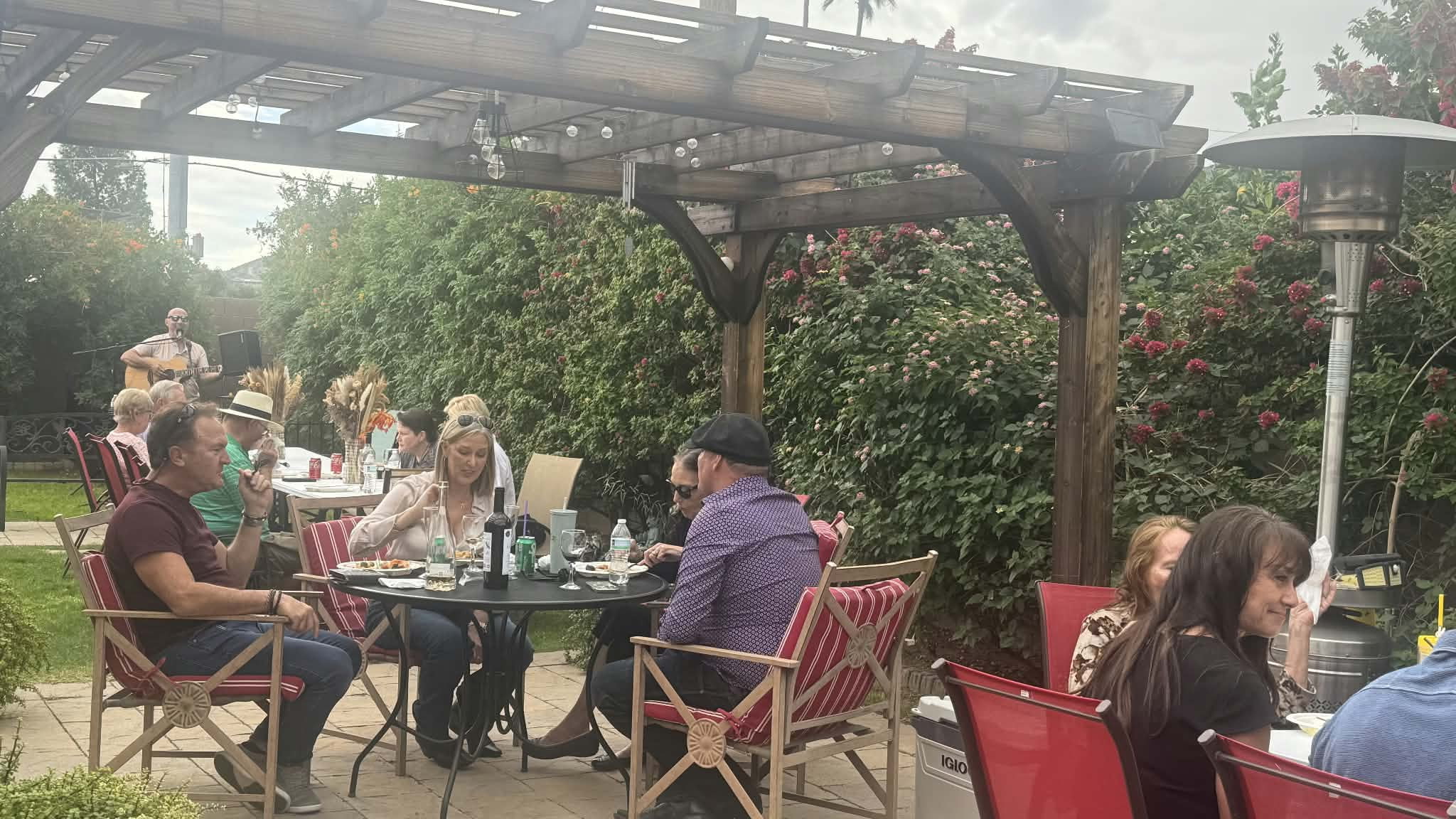 Guests sharing a meal under the garden pergola while a musician plays nearby